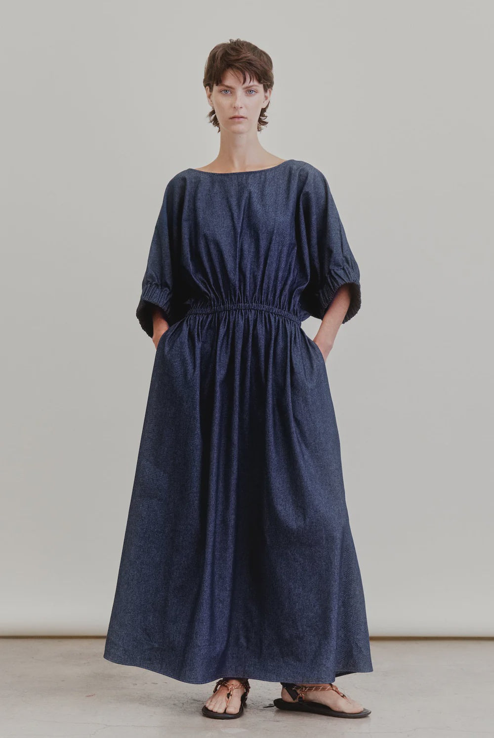 A person stands against a plain backdrop wearing Aquarius Cocktail's French 75, an ankle-length, loose-fit Japanese denim dress with a boat neck, elastic waist, short sleeves, and pockets; brown sandals complete the look.