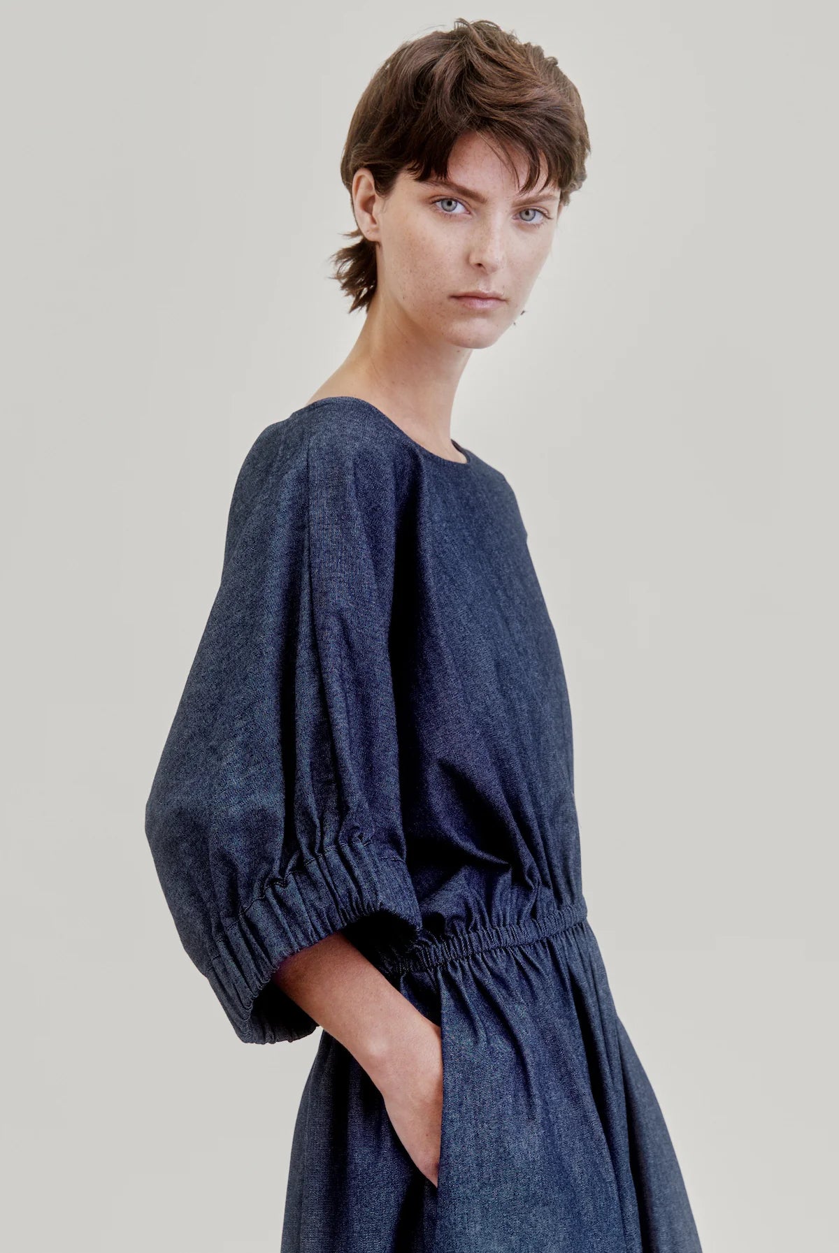 A person with short brown hair models the Aquarius Cocktail French 75 dress—a loose Japanese denim style featuring a boat neckline, puffed elbow sleeves, elastic waist, and pockets—standing against a plain light background.