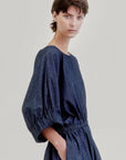A person with short brown hair models the Aquarius Cocktail French 75 dress—a loose Japanese denim style featuring a boat neckline, puffed elbow sleeves, elastic waist, and pockets—standing against a plain light background.