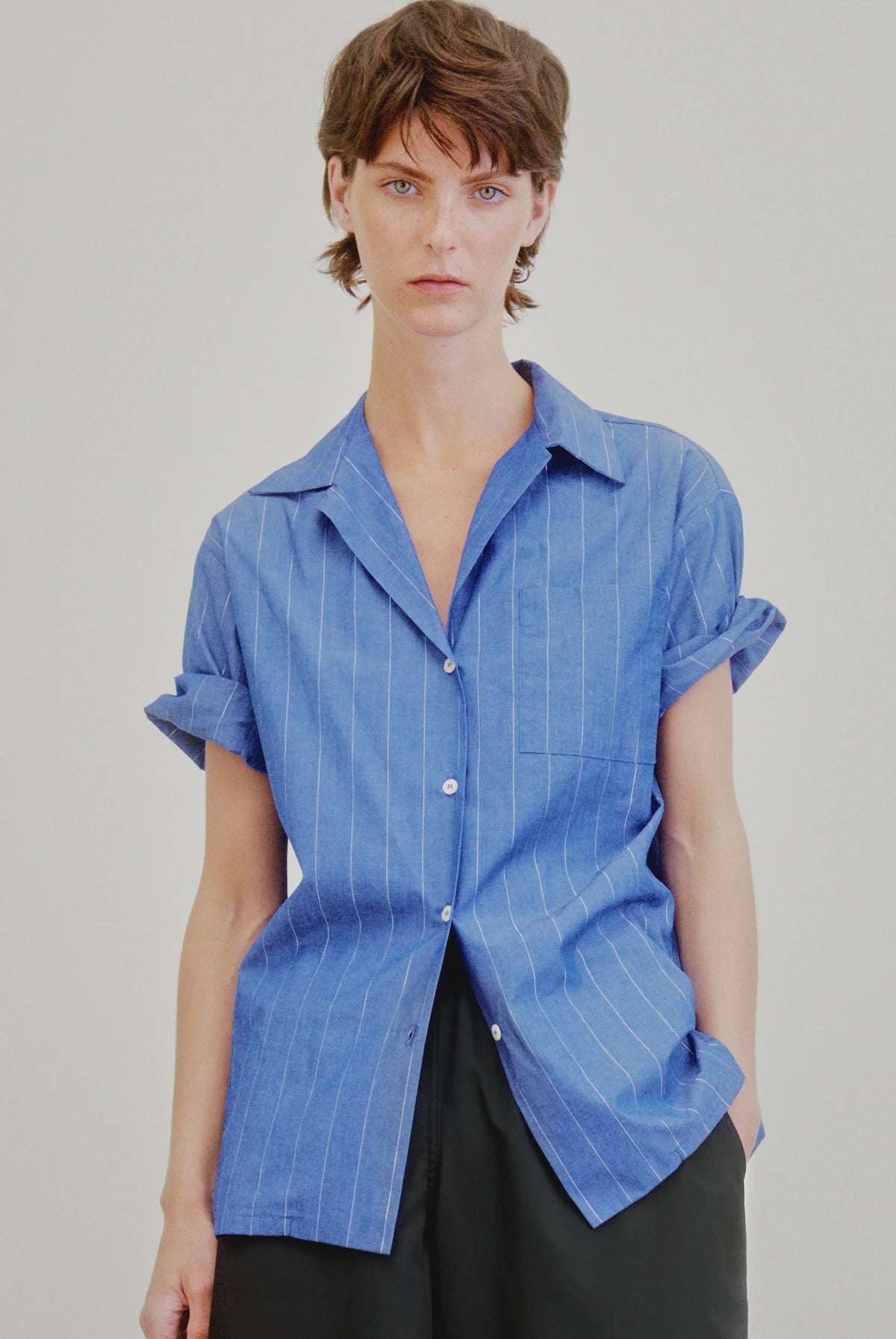 A person with short brown hair wears the Aquarius Cocktail Le Camp SS Shirt with Pocket, a blue Japanese cotton camp-style shirt featuring white pinstripes, paired with black pants against a plain light background.