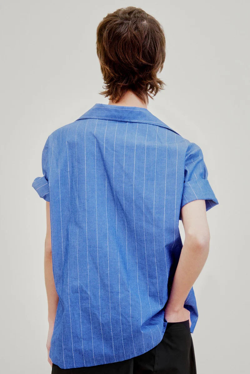 A person with short brown hair stands with their back to the camera, wearing the Aquarius Cocktail Le Camp SS Shirt with Pocket—blue Japanese cotton with thin white stripes—and black pants, one hand in their pocket, against a plain light background.