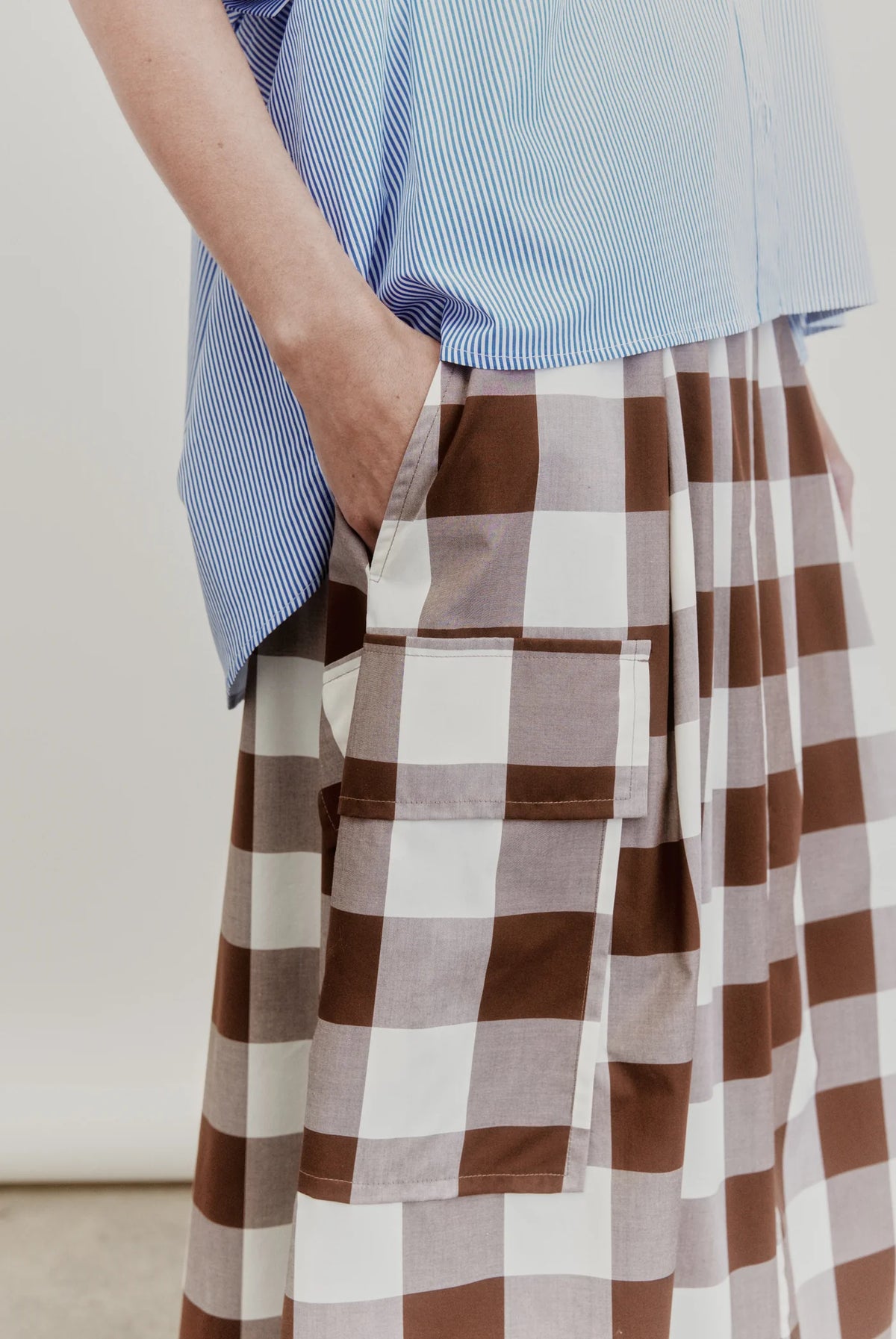 Wearing the Aquarius Cocktail Le Cargo Skirt, crafted from Japanese cotton poplin in a brown-and-white checkered pattern, a person stands with one hand tucked into one of the skirt’s oversized cargo pockets, paired with a blue striped top.