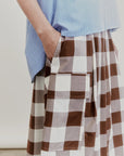 Wearing the Aquarius Cocktail Le Cargo Skirt, crafted from Japanese cotton poplin in a brown-and-white checkered pattern, a person stands with one hand tucked into one of the skirt’s oversized cargo pockets, paired with a blue striped top.