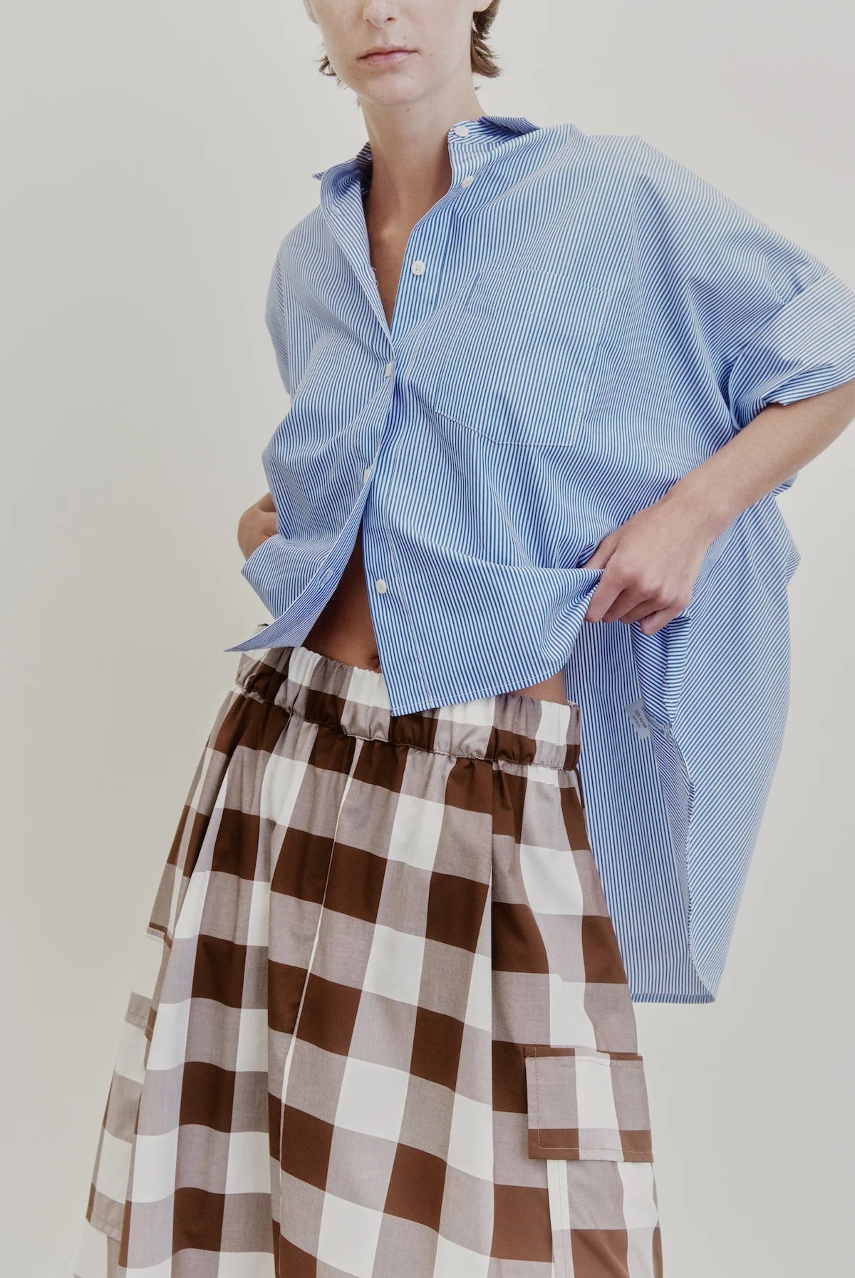 Wearing an oversized blue striped shirt crafted from Japanese cotton poplin, paired with Aquarius Cocktail’s Le Cargo Skirt—a high-waisted brown and white checkered skirt featuring an elastic waist—one hand lifts the shirt slightly. The background is plain and light-colored.