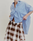 Wearing an oversized blue striped shirt crafted from Japanese cotton poplin, paired with Aquarius Cocktail’s Le Cargo Skirt—a high-waisted brown and white checkered skirt featuring an elastic waist—one hand lifts the shirt slightly. The background is plain and light-colored.