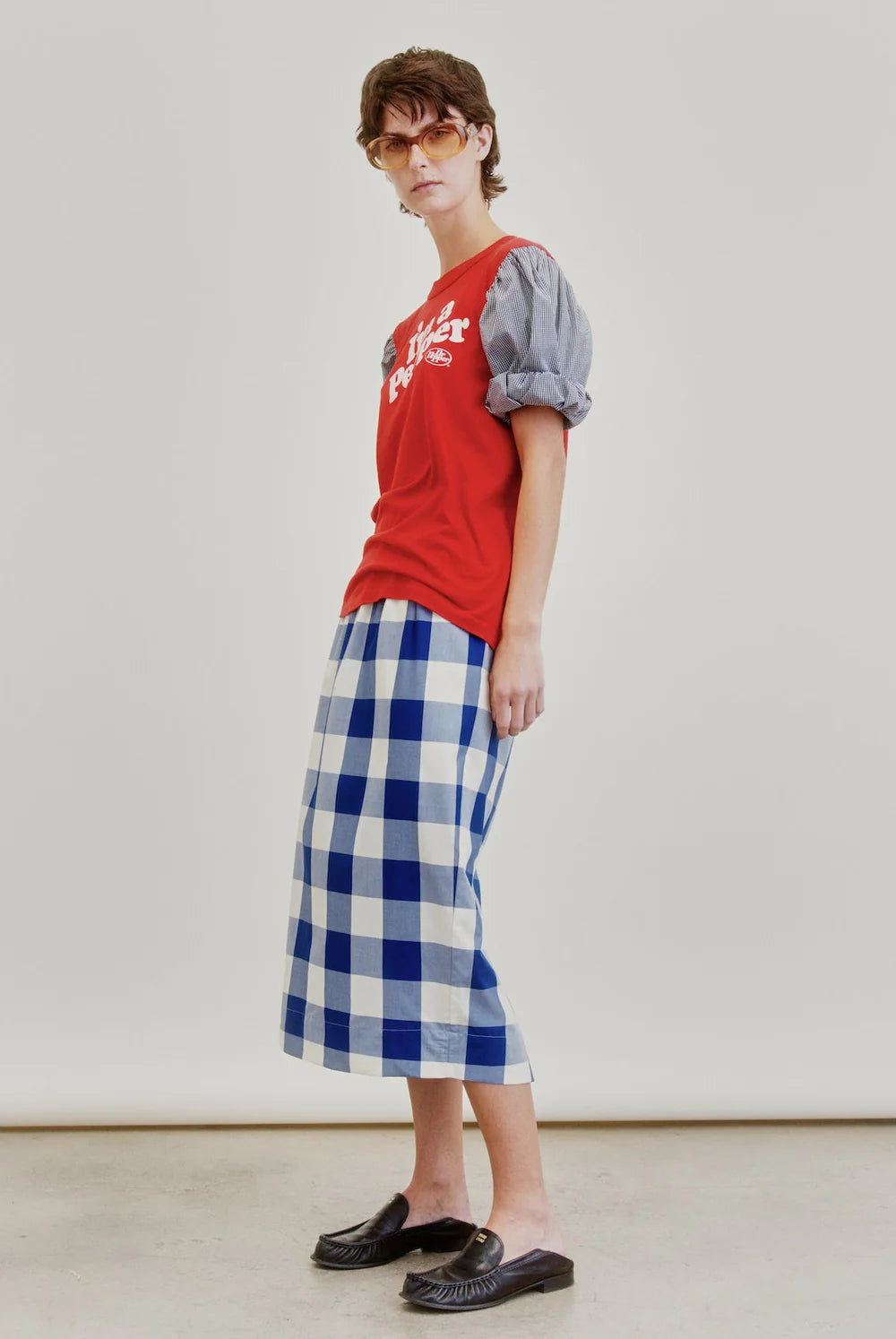 A person wearing sunglasses, a red graphic T-shirt with puffed gray sleeves, the Le Pencil Slim Skirt by Aquarius Cocktail in blue and white checkered Japanese cotton poplin, and black slip-on shoes stands against a plain, light background.
