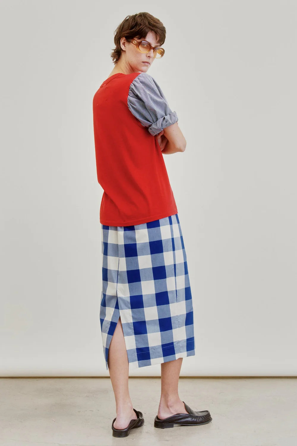 A person wearing sunglasses, a red vest layered over a blue-gray shirt, and the Aquarius Cocktail Le Pencil Slim Skirt—featuring a blue-and-white checkered pattern, side slit, and crafted from Japanese cotton poplin—paired with black sandals stands against a plain background, looking over their shoulder.