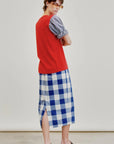 A person wearing sunglasses, a red vest layered over a blue-gray shirt, and the Aquarius Cocktail Le Pencil Slim Skirt—featuring a blue-and-white checkered pattern, side slit, and crafted from Japanese cotton poplin—paired with black sandals stands against a plain background, looking over their shoulder.