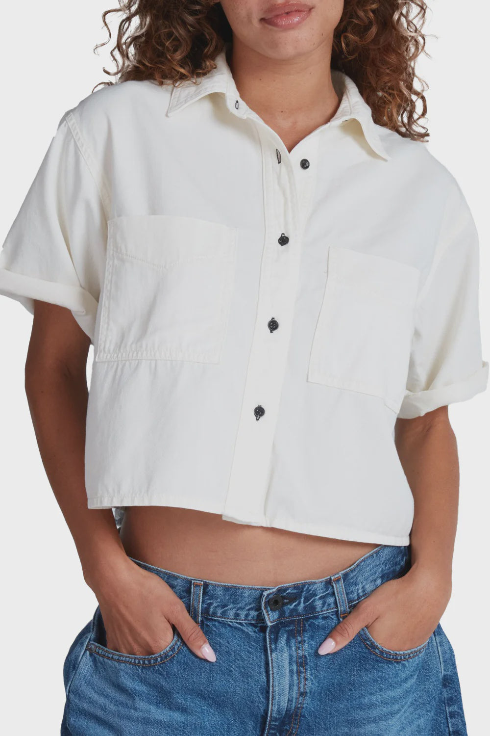 Wearing the SS SHASHIKO, a cropped boxy-fit white short-sleeve button-up with two chest pockets, paired with blue jeans. The slightly cropped shirt is styled casually with hands in pockets.