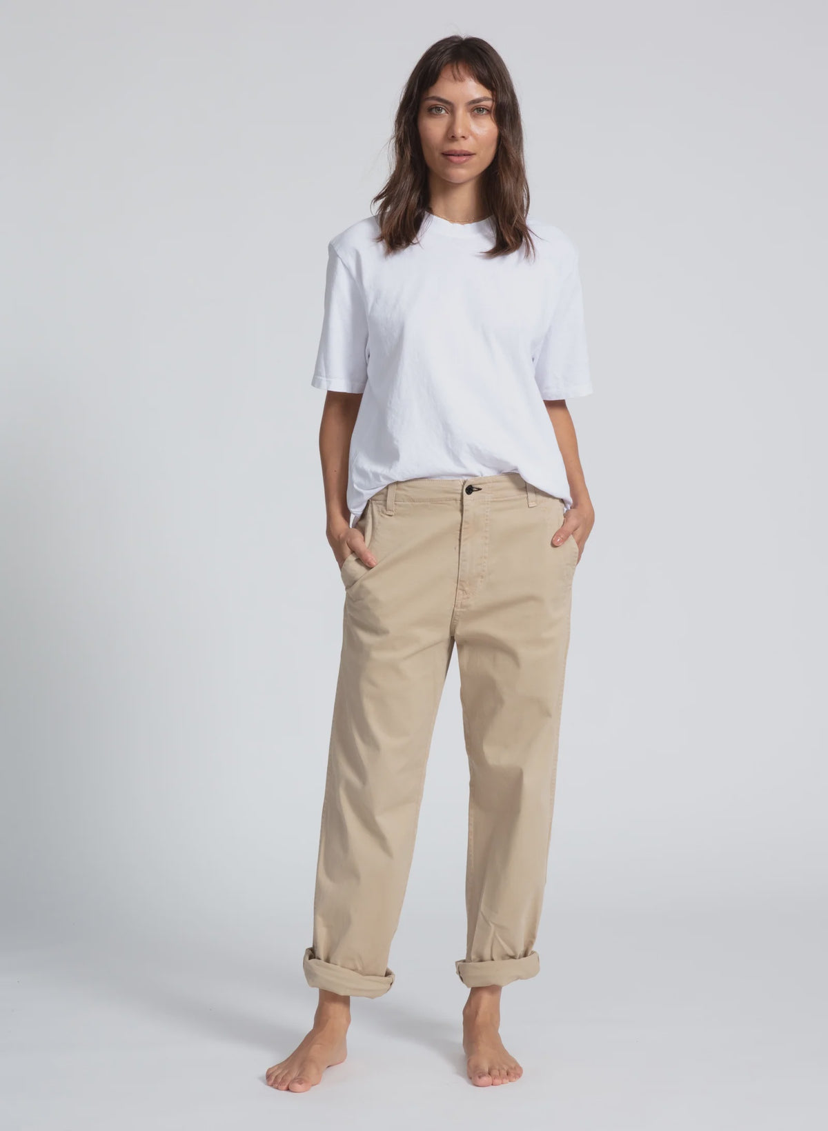 A woman stands barefoot against a plain backdrop, wearing a loose-fitting white T-shirt and ASKK's beige rolled-up chino twill pants. Her hands are in her pockets, and she is looking directly at the camera with a neutral expression.
