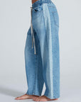 A person is wearing the ASKK Cancun Pant: light blue, wide-leg Japanese denim jeans with an elastic waistband, drawstring, and two-tone outer leg panels in a light Berkeley wash. The person is barefoot.
