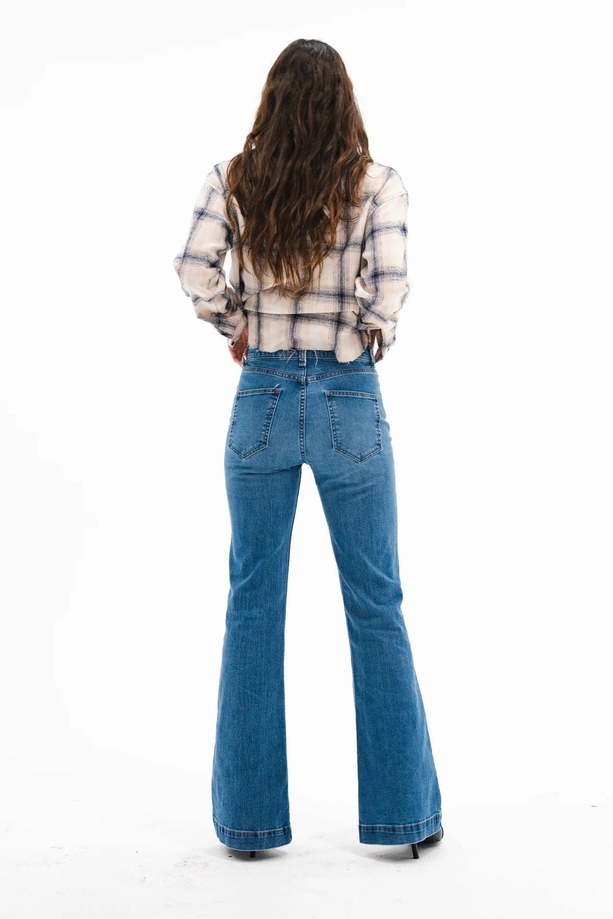 A woman with long wavy brown hair, wearing the ASKK Crosby Flannel in Japanese shirting fabric and high-waisted blue flared jeans, stands facing away against a plain white background, styled with black shoes.