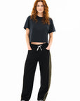 A woman with long curly red hair stands facing forward, wearing a loose dark t-shirt and ASKK’s Cancun Pant—wide leg, cropped drawstring black pants with an olive green side panel—plus black-and-white sneakers, hands in her pockets.