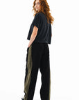 A person with long curly hair wears a loose black T-shirt, ASKK Cancun Pant in black with olive side stripes, and black sneakers, standing and looking over their shoulder against a white background.
