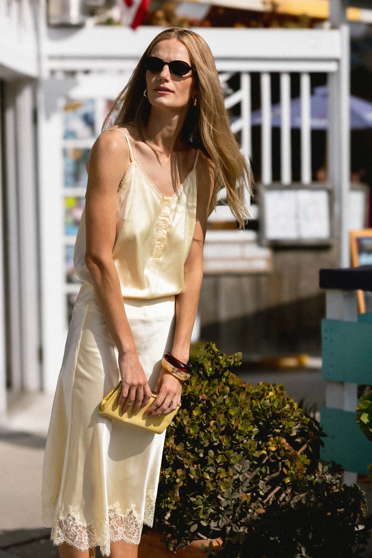 A woman in THE SHIRT's The Slip Skirt, sunglasses, and holding a yellow clutch stands outdoors by greenery and a white fence, sunlight highlighting her outfit.