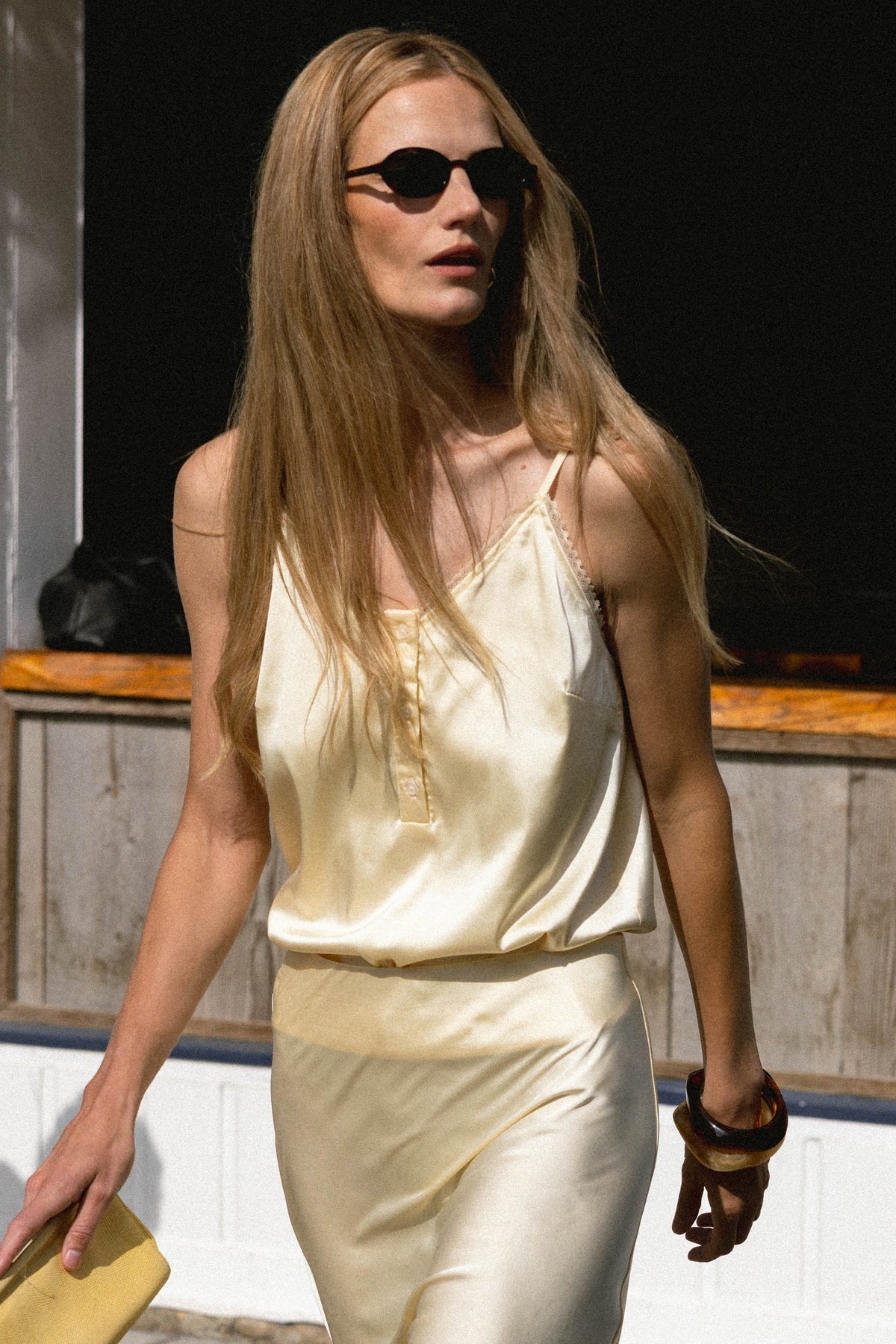 A woman with long blonde hair and black sunglasses walks outdoors, wearing THE SHIRT’s The Cami Shirt—a light yellow sleeveless top with a No Gape button—paired with a yellow clutch and chunky bracelet.