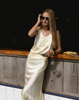 A woman in THE SHIRT’s The Slip Skirt, a cream poly satin style with lace trim, stands by a wooden counter holding sunglasses and a phone, her long hair cascading as she strikes a stylish, relaxed pose.