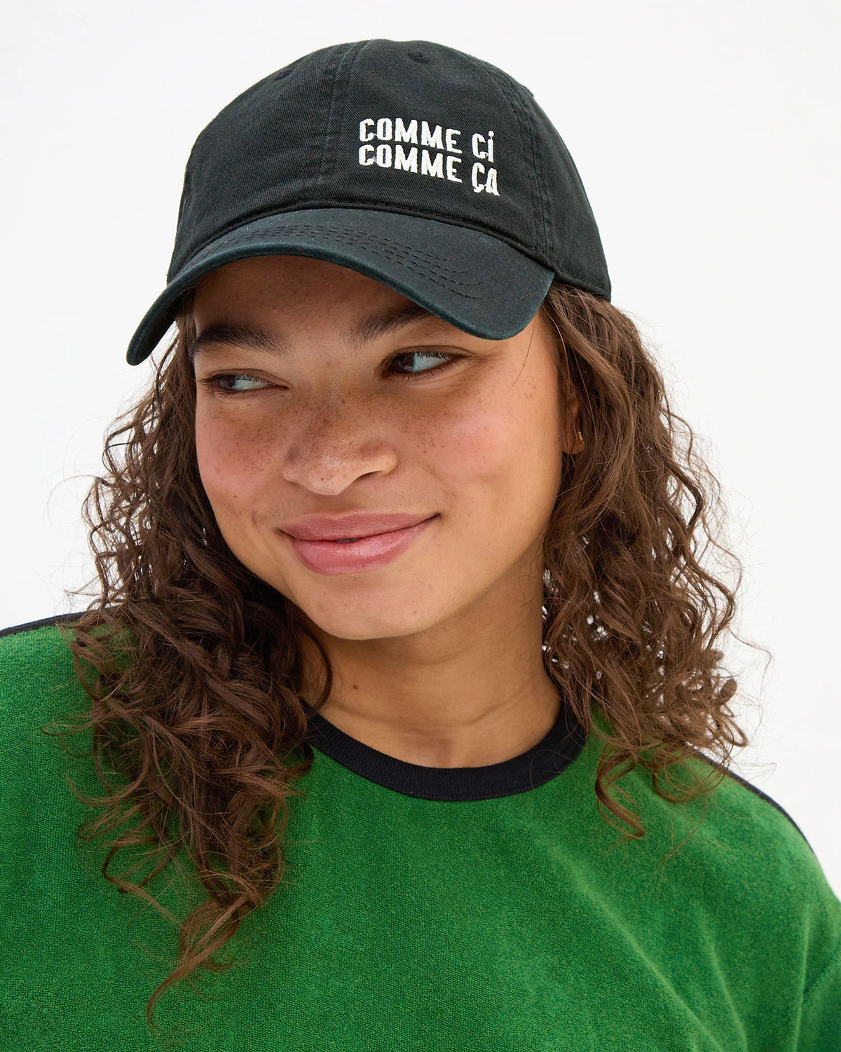 A person with curly brown hair, in a green shirt and a Clare Vivier "Comme Ci Comme Ca" Baseball Hat, looks slightly to the side and smiles gently. The black cotton hat has an adjustable sliding metal closure for a one-size-fits-most fit.