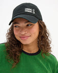 A person with curly brown hair, in a green shirt and a Clare Vivier "Comme Ci Comme Ca" Baseball Hat, looks slightly to the side and smiles gently. The black cotton hat has an adjustable sliding metal closure for a one-size-fits-most fit.
