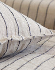 Close-up of beige Blake Big Pillow 28x36 by POM POM, featuring black vertical stripes that accentuate the hand-loomed texture and woven pattern. Part of the same style, another similarly textured surface is interwoven in the background.