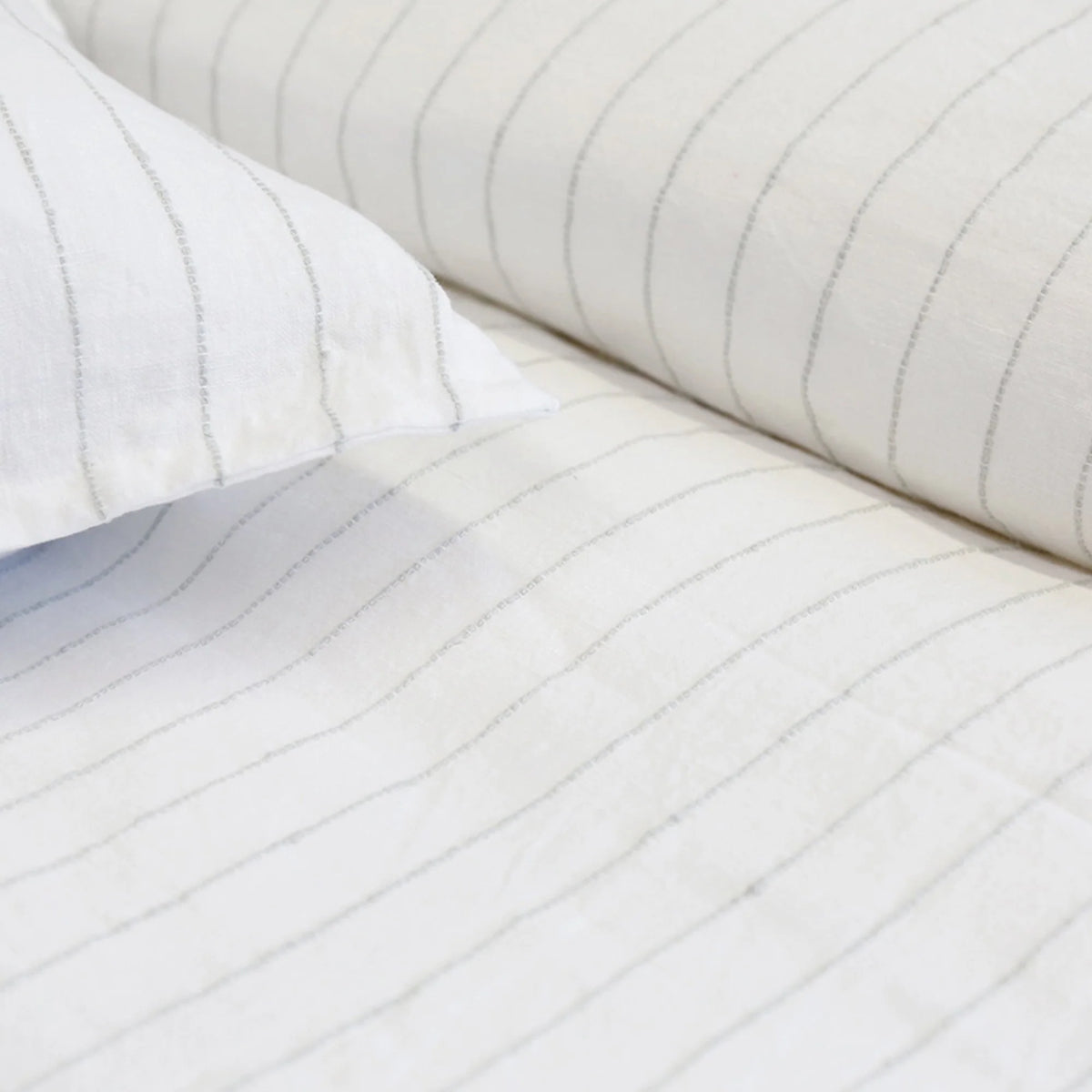 Close-up of white bed sheets and pillowcases featuring the Blake Big Pillow 28x36 from POM POM, showcasing light gray interwoven stripes. The fabric appears soft and neatly arranged, creating a clean and minimalist look.