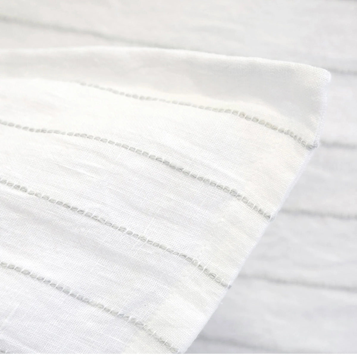 A close-up of the Blake Big Pillow 28x36 by POM POM reveals a white fabric adorned with subtle, sophisticated gray stripes, highlighting its hand-loomed texture and pattern. The material appears soft and lightweight, elegantly folded at an angle.