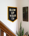 Black “Blow Up Your TV” camp flag by Bungalow Furniture & Accessories displayed above a plant in a cozy setting.