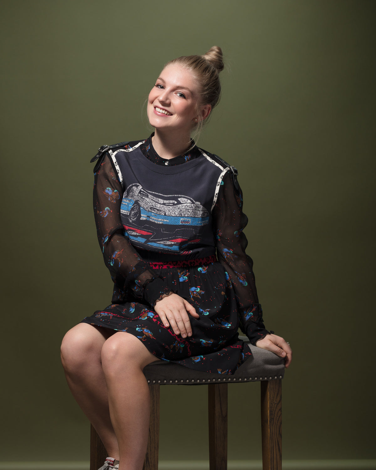 A smiling person with light hair tied up in a bun sits on a stool against a plain olive green background. The individual is wearing a black sheer long-sleeve dress adorned with a colorful design, including a car graphic on the front—a stylish choice that perfectly captures the spirit of Scottsdale. Their hands rest on their lap.