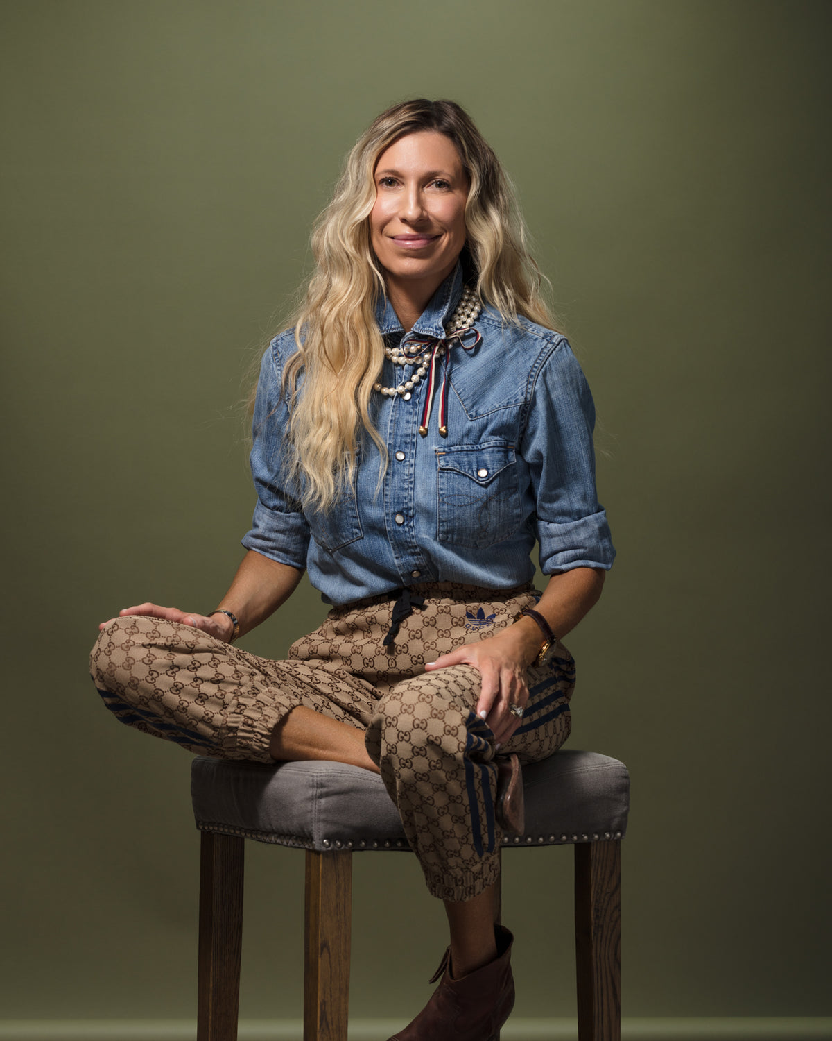A woman with long wavy hair sits on a high stool against a green background. She is wearing a blue denim shirt, patterned trousers, and brown ankle boots. She has one leg crossed over the other and is smiling gently, showcasing her Scottsdale style with layered necklaces.