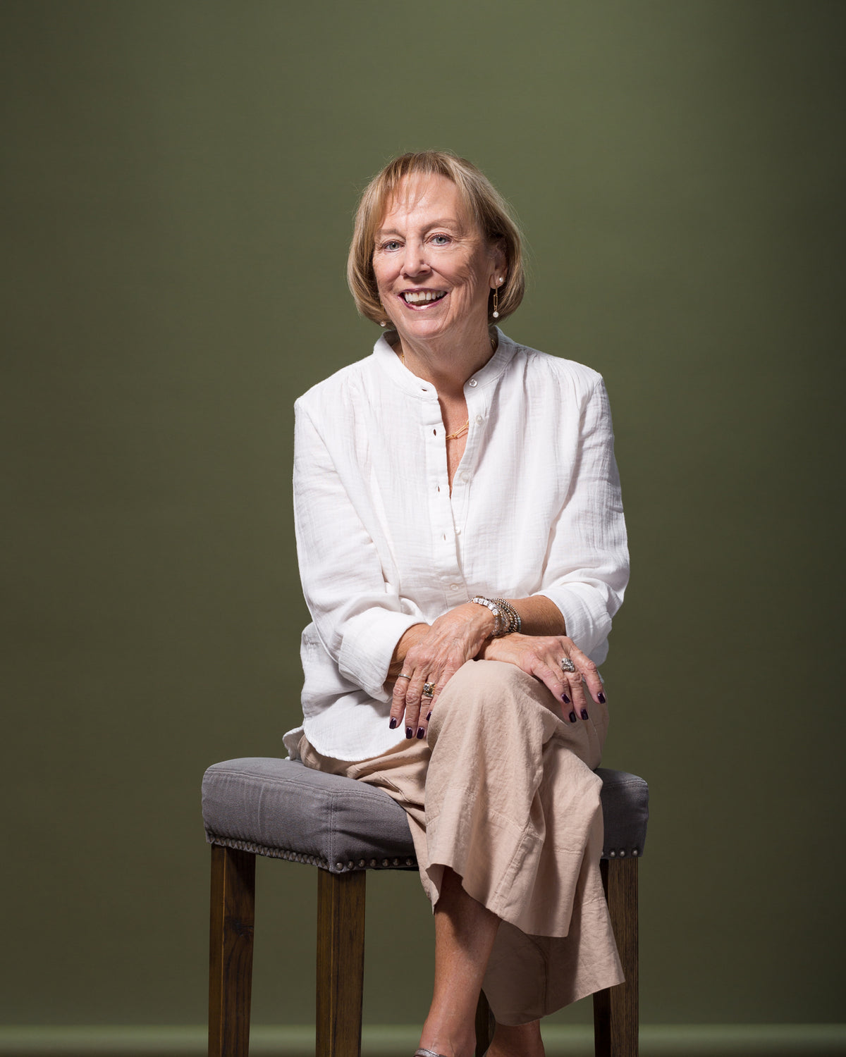 A woman with short blonde hair, dressed in a stylish white blouse and beige pants, sits on a high stool against a plain, dark green background. She is smiling and has her hands crossed on her lap, exuding an effortless Scottsdale charm.