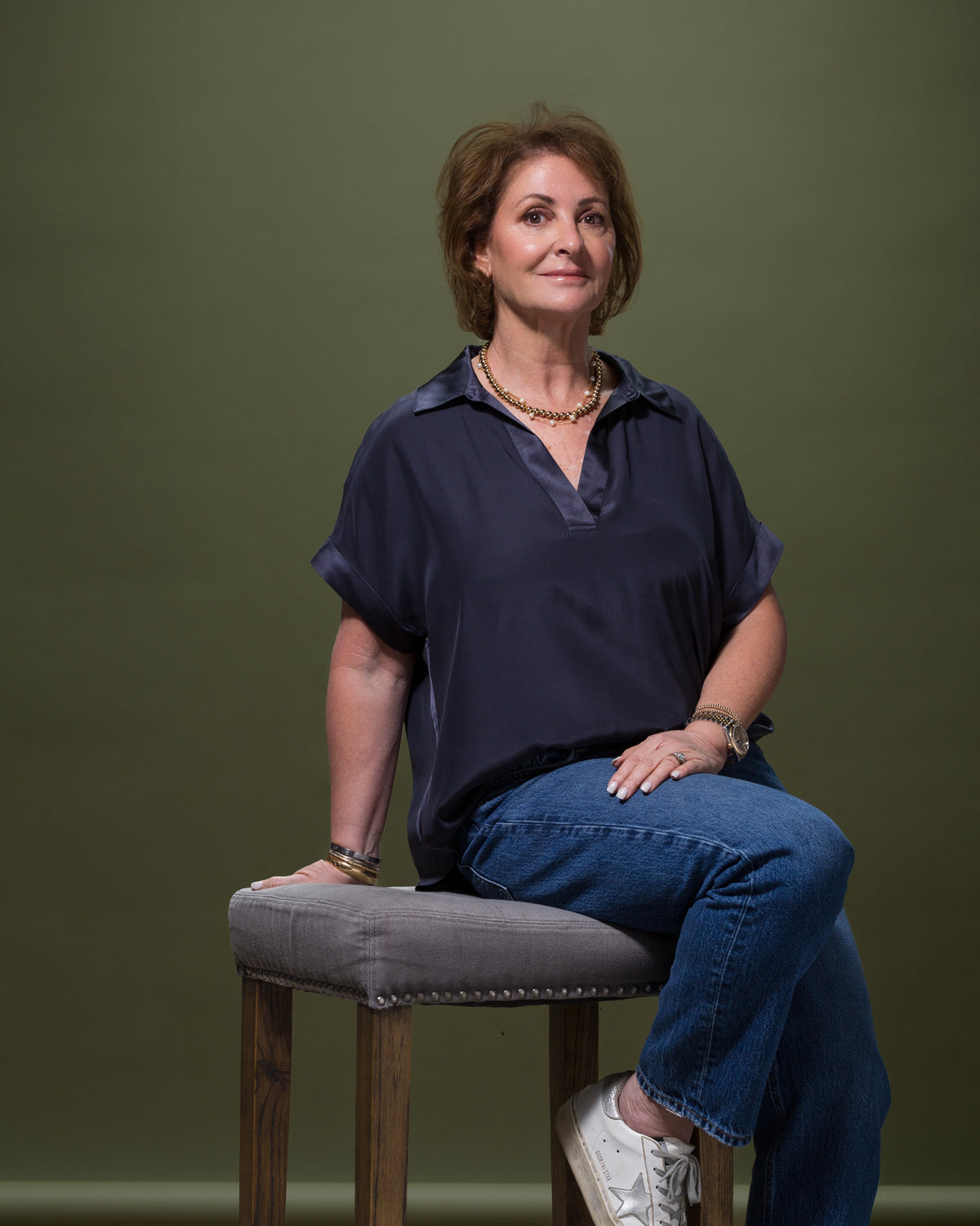 A woman with short brown hair is seated on a tall stool in a Scottsdale bungalow with a green backdrop. She is wearing a dark blue blouse, blue jeans, white sneakers, and a gold necklace that complements her stylish look. Her hands rest on her lap and the stool as she maintains a neutral expression.