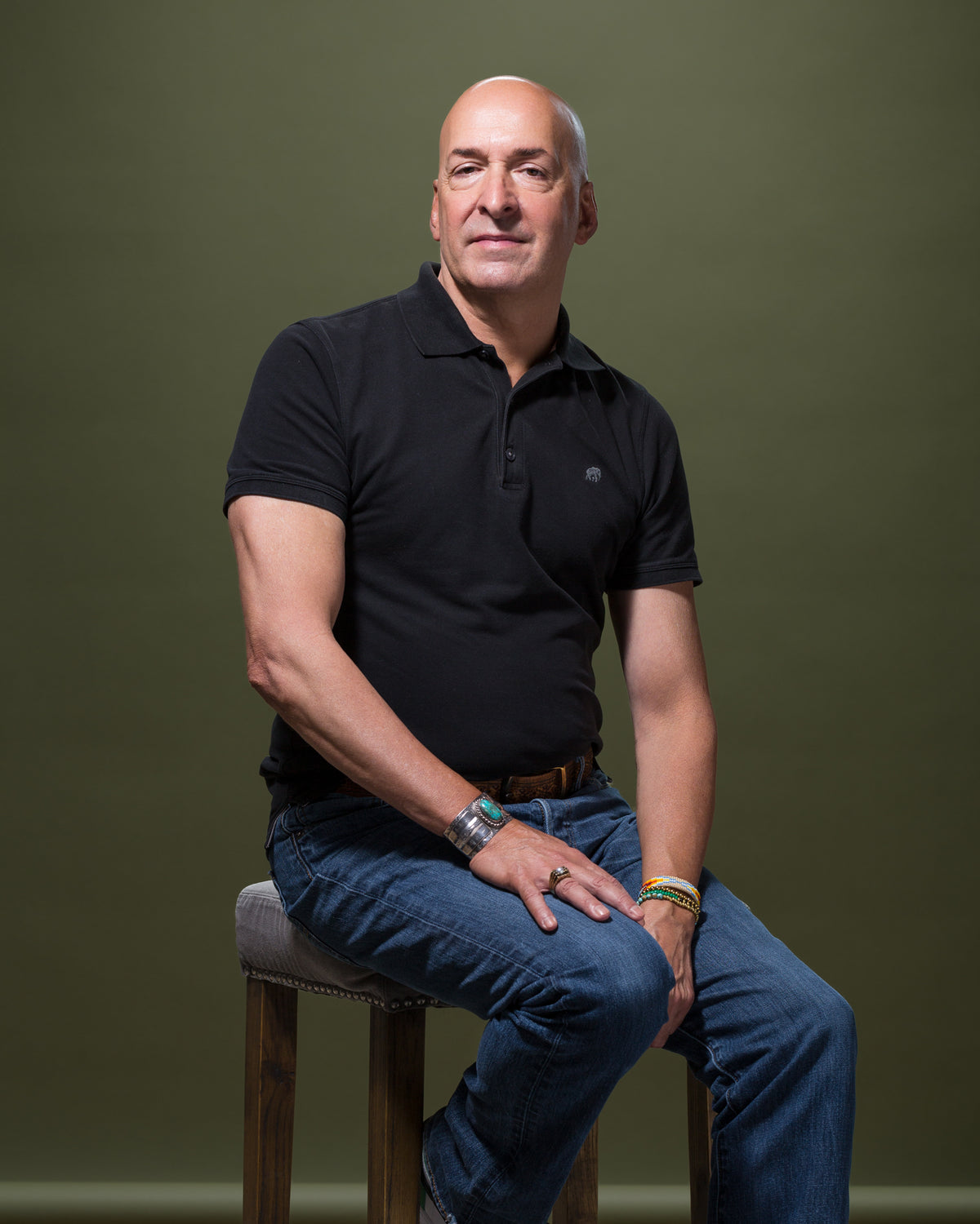 A man wearing a black polo shirt and blue jeans sits on a wooden stool against a plain, olive-green background. With the relaxed style of someone lounging in a Scottsdale bungalow, he rests one hand on his thigh and the other on the stool.