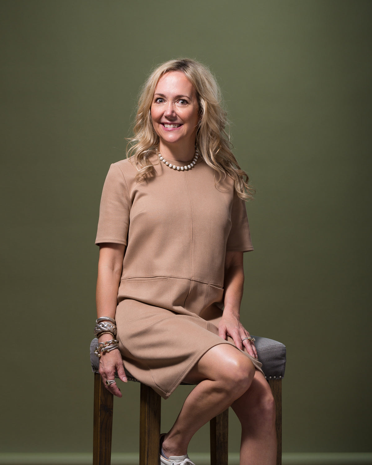 Tan dress styled on a woman sitting casually on a wooden stool with a green backdrop, Bungalow Furniture & Accessories.