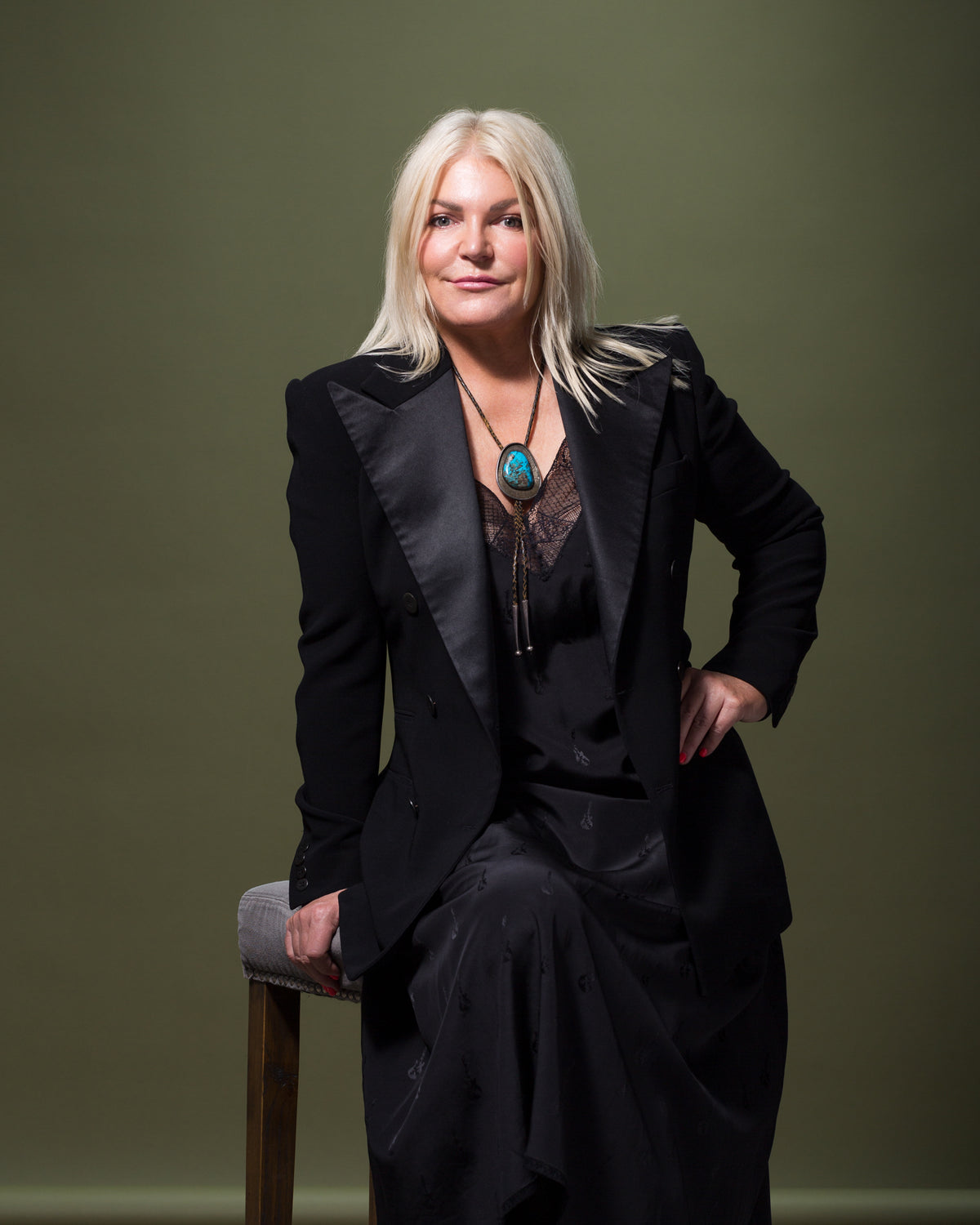 A person with long blonde hair is seated on a stool against a muted green background reminiscent of a Scottsdale bungalow. They are wearing a black suit jacket over a lacy black dress and a turquoise pendant necklace. The person is smiling and has a relaxed posture with one hand resting on the stool, exuding effortless style.