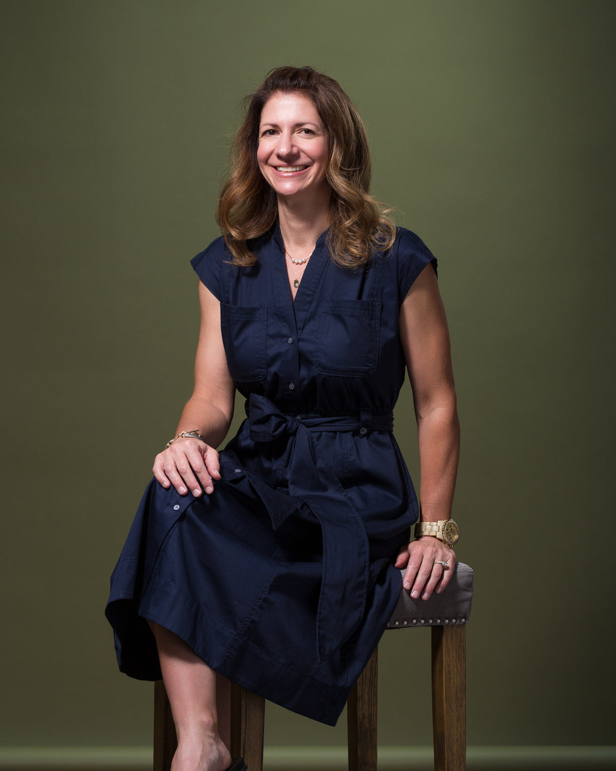 A woman with long, wavy hair is sitting on a wooden stool against a plain, olive-green background. She is wearing a dark blue, short-sleeve button-up dress with a tied waist. She is smiling and has her hands resting on her lap. Her style is complemented by a gold watch and bracelet, radiating effortless elegance.