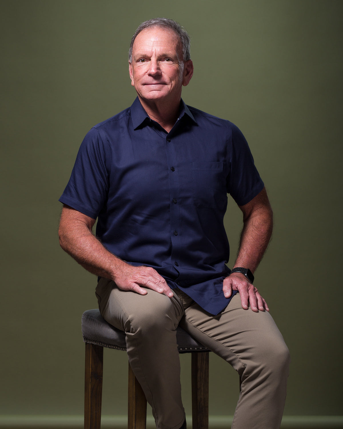 A man with short gray hair and a navy blue short-sleeve button-up shirt sits on a stool against a plain green background, evoking the timeless style of an old Scottsdale bungalow. He has a neutral expression and is wearing tan pants. His hands rest on his knees.