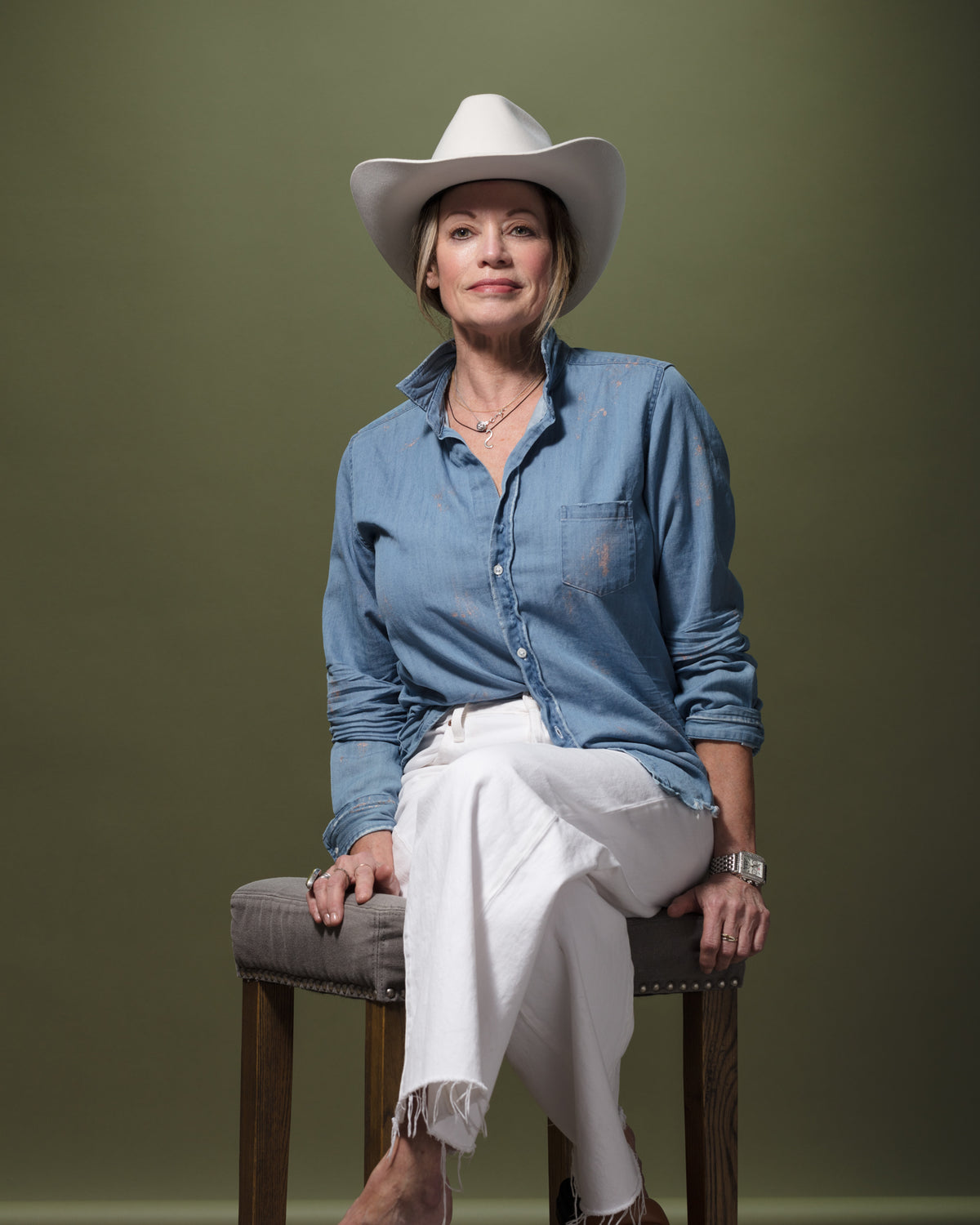 Bungalow Stylist, Suzanne, with a confident expression, wearing a white cowboy hat, button-down denim shirt, and white pants, sits on a stool against a green background. Their legs are crossed, and they have one hand resting on their knee.