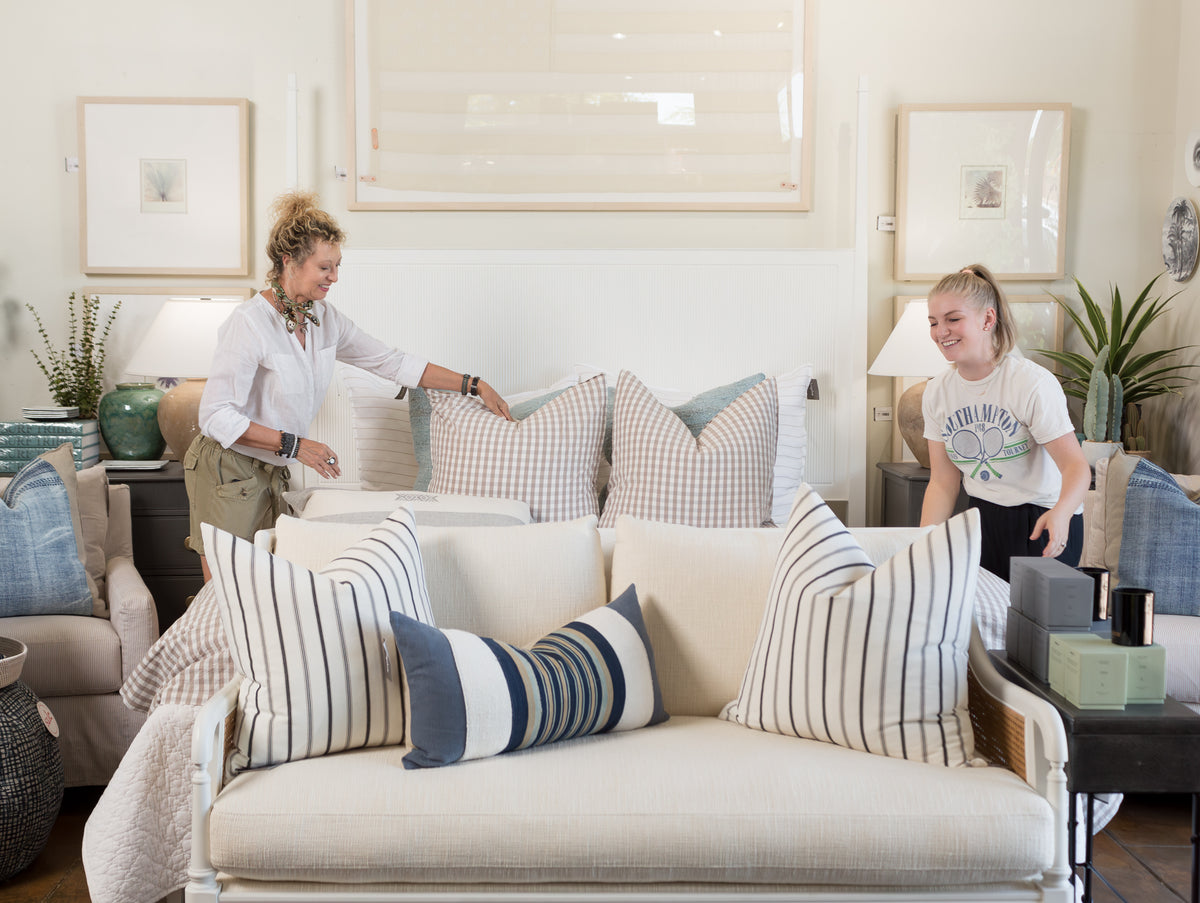Two women are arranging pillows in a light and airy Scottsdale bungalow living room. One woman is on the left, wearing a white shirt and green pants, while the other woman on the right wears a t-shirt and has her hair in a ponytail. The room has a cozy, neutral color palette with blue accents, exuding classic style.