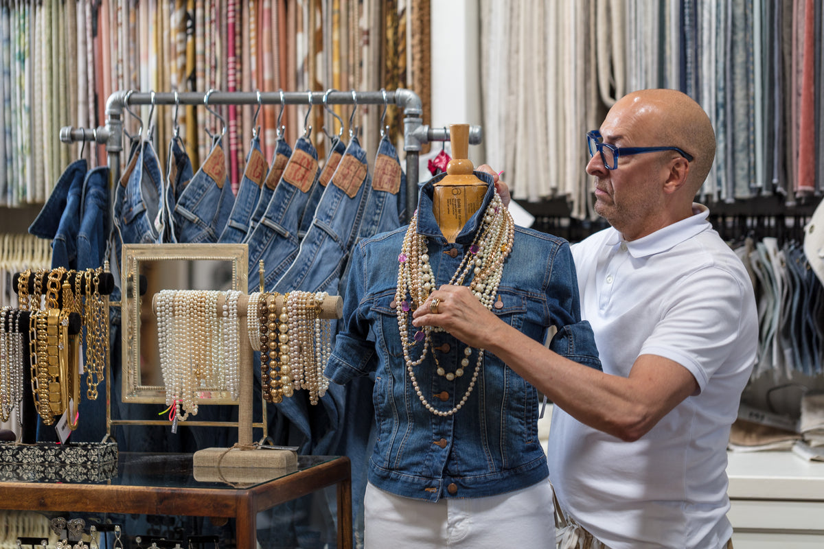 A bald man with glasses and a white shirt is arranging necklaces on a denim jacket displayed on a mannequin. Next to him, there's a rack with more jackets and a stand showcasing various necklaces and bracelets. The stylish setup contrasts beautifully against the background of fabric rolls in this Scottsdale bungalow.