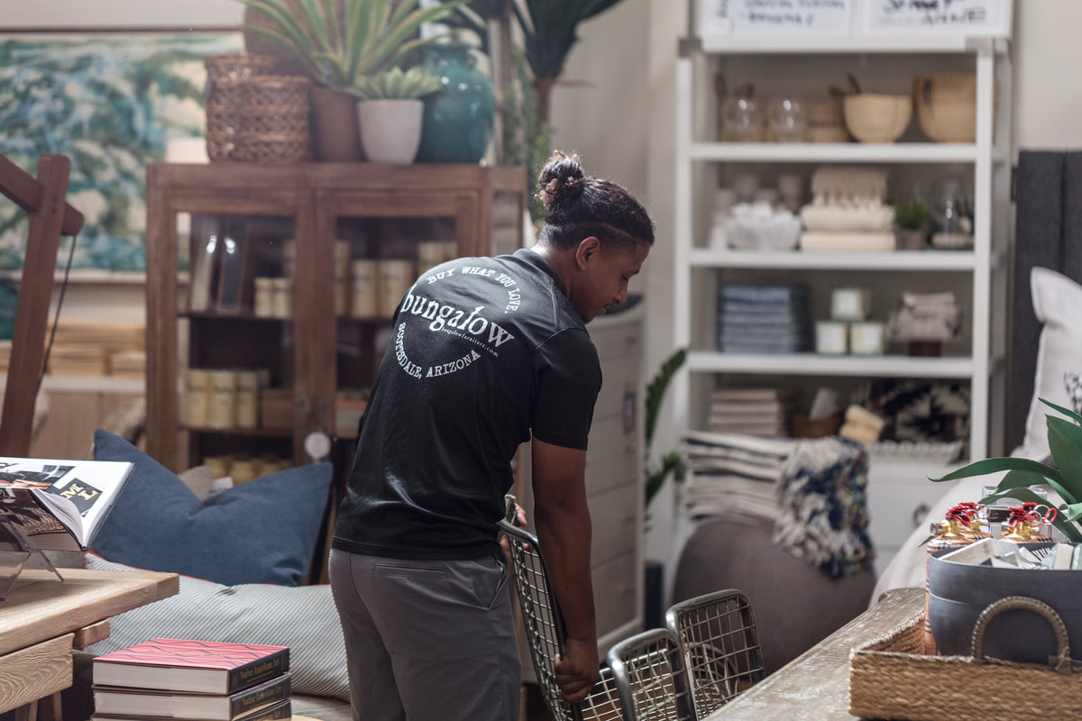 A person is arranging furniture and decor items in a stylish Scottsdale store. They are wearing a black shirt and facing away from the camera. The store's interior has shelves filled with various home decor items, including baskets, plants, and textiles that evoke a charming bungalow feel.