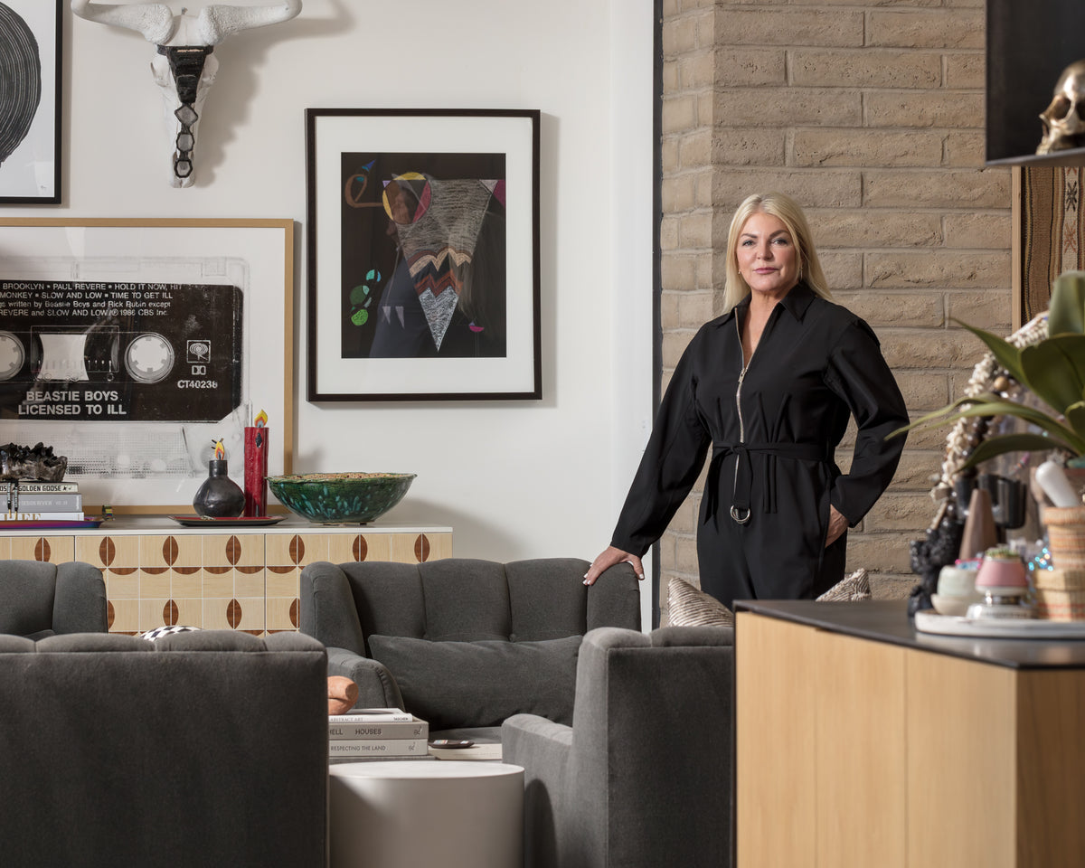 A woman with long blonde hair stands in a modern living room of a stylish Scottsdale bungalow, wearing a black outfit. The room features grey furniture, eclectic artwork, and various decor items, including a framed record and posters on the walls. A brick wall is partially visible in the background.