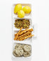 A rectangular white Montes Doggett 5455 Appetizer Platform No. 455 contains yellow lemons in the top section, shelled pistachios beneath them, twisted handmade breadsticks in the third section, and nests of dried pasta in the bottom section.