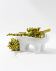 Montes Doggett's 4846 Bowl No.846, a white high-fired ceramic dish with a unique four-legged stand, elegantly holds a bunch of green bananas. Crafted in Peru, the bowl and fruit create a striking contrast against the plain white background, achieving a minimalistic and modern aesthetic.