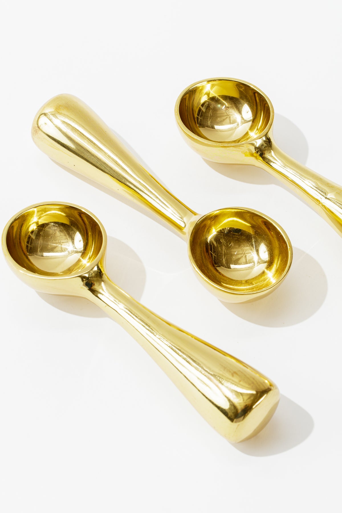 Three shiny, 100% food-safe Brass Dessert Scoops from Sir/Madam with a modern, rounded, and sleek handle design laid on a white background. The scoops are positioned at different angles, showcasing their reflective surface and making them a perfect addition to any kitchenalia collection.
