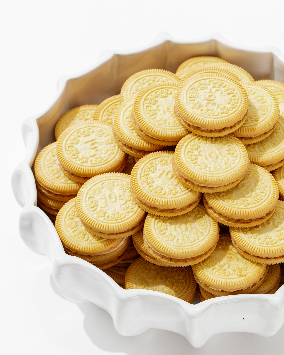 Bowl No. 727 by Montes Doggett is a high-fired, white ceramic bowl made in Peru, overflowing with a large number of golden sandwich cookies. Each cookie boasts a patterned design and cream filling, presented against a plain white background.