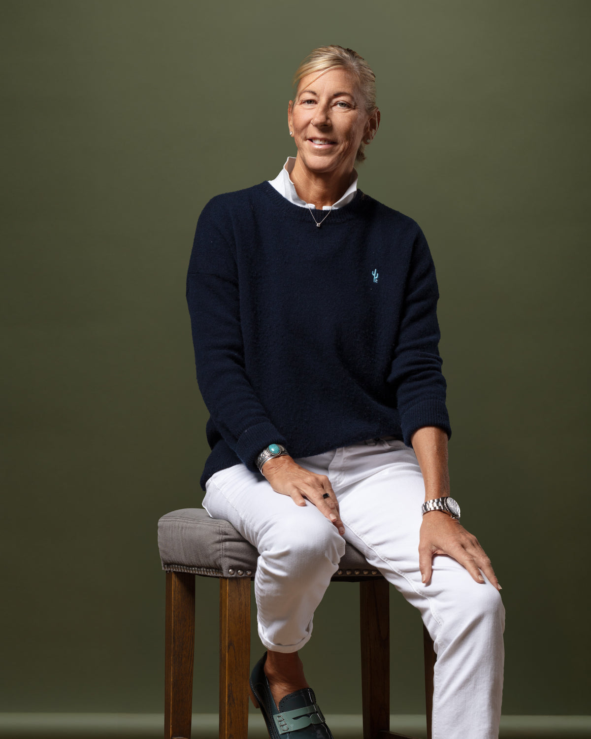 Navy sweater and white pants from Bungalow Furniture worn by model seated casually on a stool with olive backdrop.