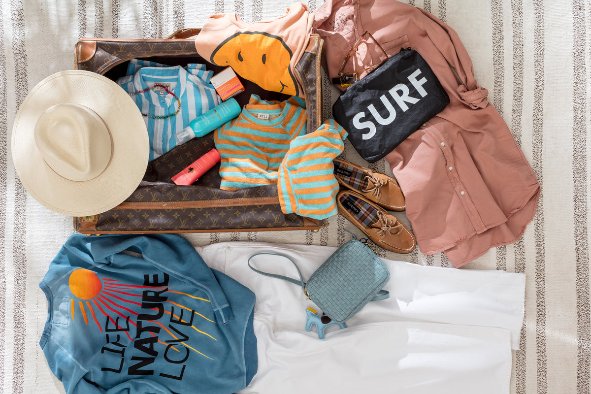 Striped shirts and summer essentials in an open suitcase, styled on striped fabric for Bungalow’s travel collection.