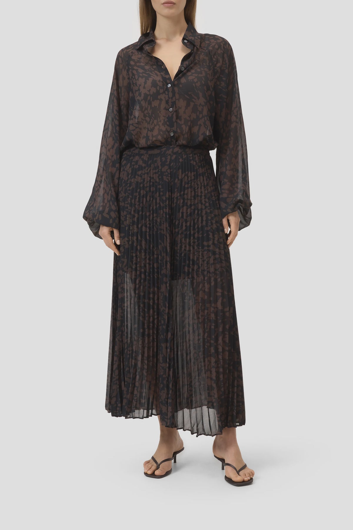 Closed pleated skirt and sheer dark patterned blouse, styled with black sandals; minimal backdrop, Bungalow Furniture.
