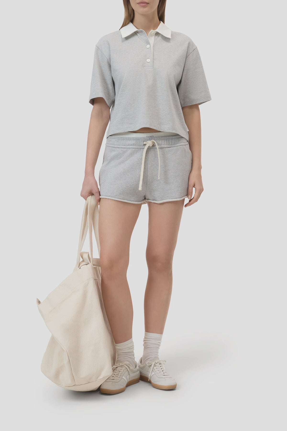 A woman wears the CLOSED Cropped Rugby Shirt in light gray with matching drawstring shorts, white sneakers, socks, and an off-white tote bag. The look features a relaxed silhouette against a plain, light-colored background.