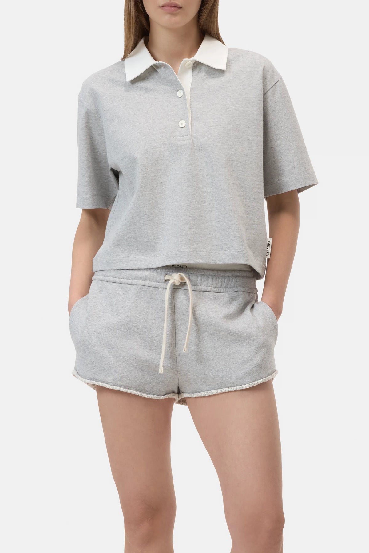 A woman wears the CLOSED Cropped Rugby Shirt in light gray with a white collar and buttons, paired with matching drawstring shorts, standing hands-in-pockets against a plain background.